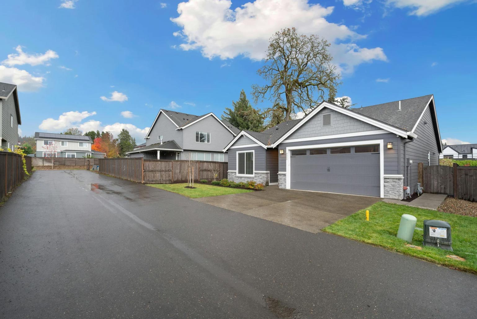 Gray single-story house with a two-car garage and driveway on a wet suburban street under a blue sky.