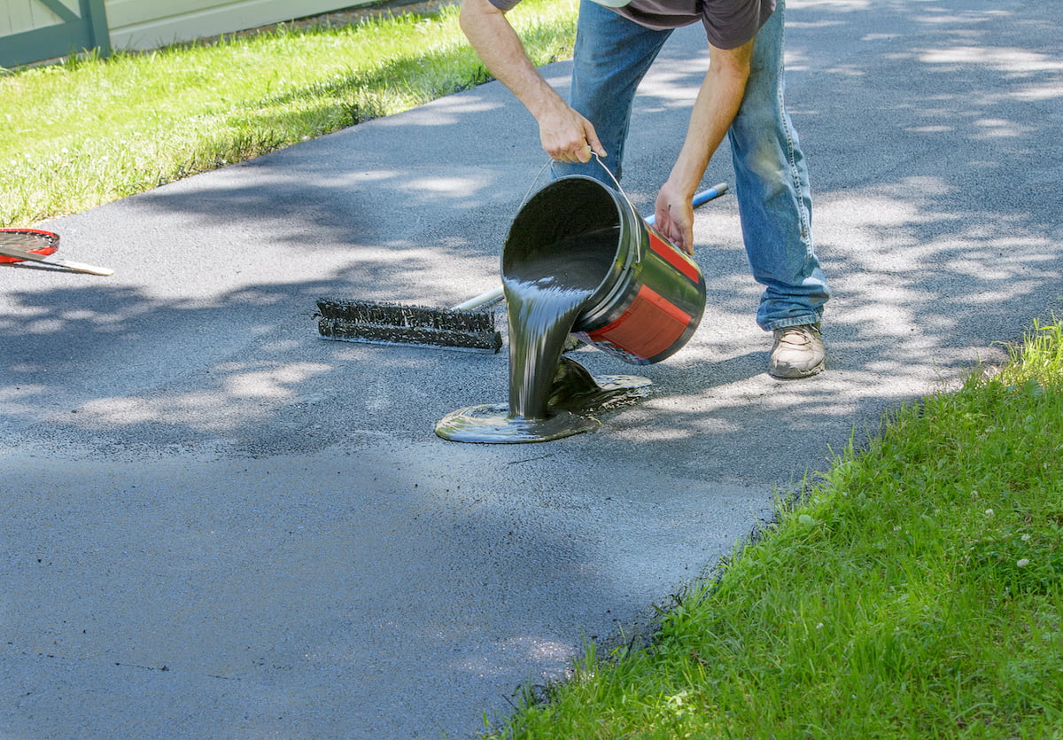 maintenance-repair-driveway Person pouring black sealant from a bucket onto a driveway, preparing to spread it with a tool.