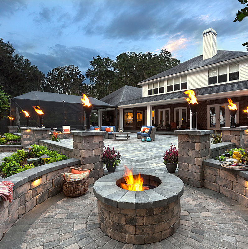 Spacious backyard patio with a fire pit, torches, seating, plants, and a large house in the background.
