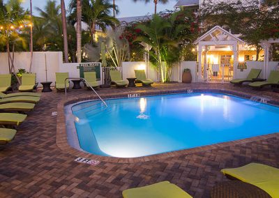 A small outdoor pool surrounded by green lounge chairs and palm trees, lit up in the evening.
