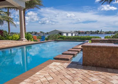 Luxurious backyard pool with steps, palm trees, and a waterfront view under a sunny blue sky.