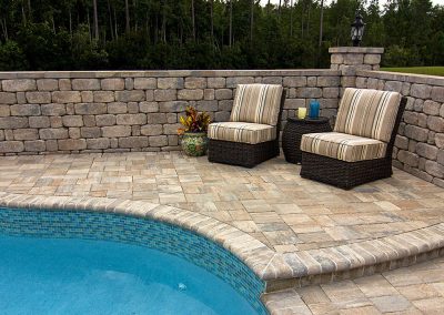 Two striped chairs and a small table sit on a stone patio beside a pool, with a stone wall in the background.