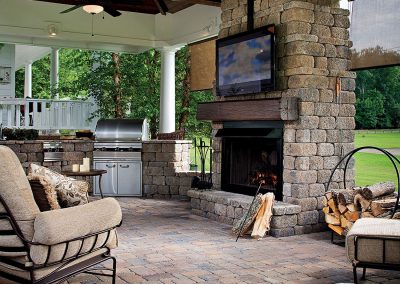 Covered patio with stone fireplace, TV, grill, cushioned seating, and stacked firewood, overlooking a lawn.