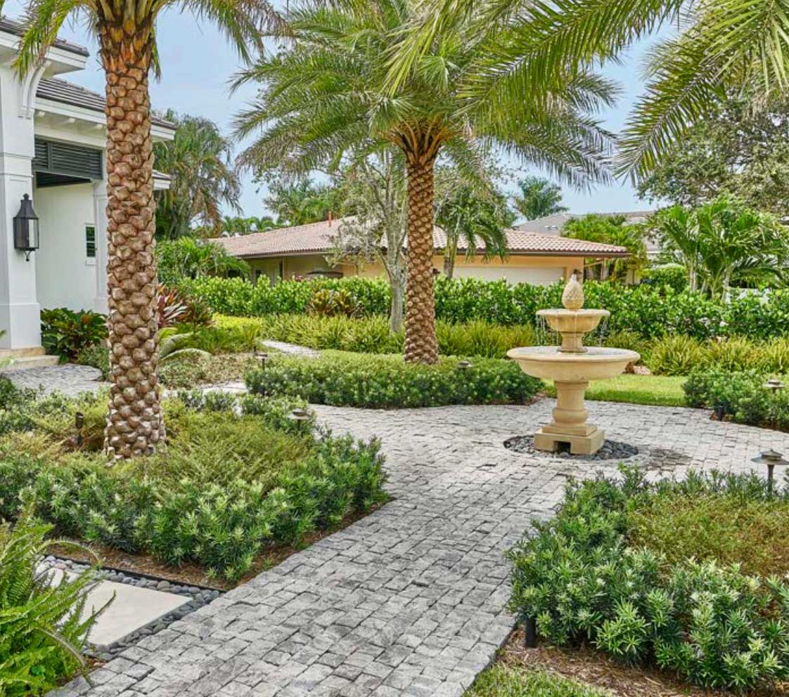 Stone path with lush greenery, palm trees, and a central fountain in a landscaped garden.