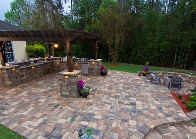 Spacious patio with stone pavers, pergola kitchen, curved seating wall, and potted flowers, backed by trees.