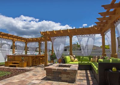 Wood pergola with white curtains, green patio furniture, and a fire pit on a stone patio under a blue sky.