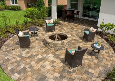A circular patio with four chairs and tables surrounds a stone fire pit in a landscaped backyard.