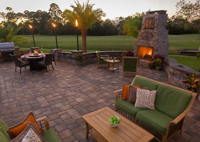 Patio with green-cushioned seating, dining table, fireplace, grill, torches, and lawn at sunset.
