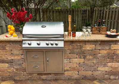Outdoor brick kitchen with a stainless steel grill, drinks, plates, and fruit on a granite countertop.