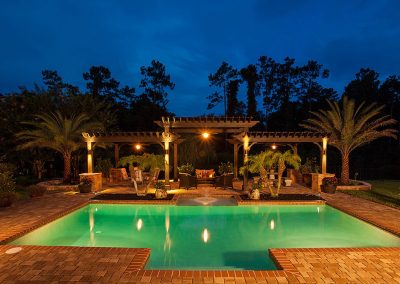 A lit outdoor pool with a pergola, patio seating, palm trees, and brick pavement at dusk.