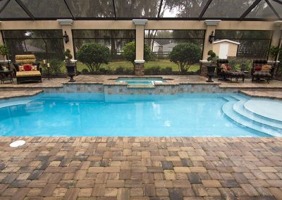 A screened-in backyard pool with a hot tub, surrounded by stone pavers and outdoor seating.