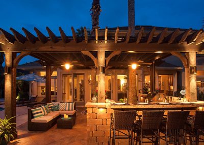 Outdoor pergola with warm lights, bar seating, and lounge area by a pool at dusk.