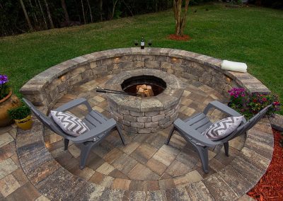 Two chairs with cushions by a circular stone fire pit, surrounded by flowers and a grassy yard.