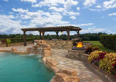 Stone pool with waterfall, fire pit, pergola, lush plants, and hedge under a partly cloudy sky.