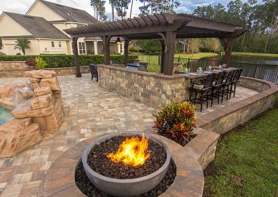 Stone patio with fire pit, outdoor kitchen, bar seating, pool, and a covered pergola near a pond and trees.