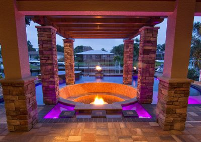 Outdoor patio with stone pillars, fire pit, and purple pool lighting in a backyard at dusk.