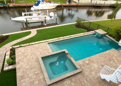 A backyard with a pool, hot tub, green lawn, and boats docked by a canal.