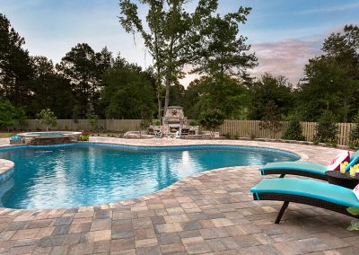 Curved backyard pool with lounge chairs, stone patio, hot tub, and trees in the background.