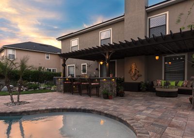 Backyard patio with pergola, bar seating, pool, cushioned seating area, and stone paving at sunset.