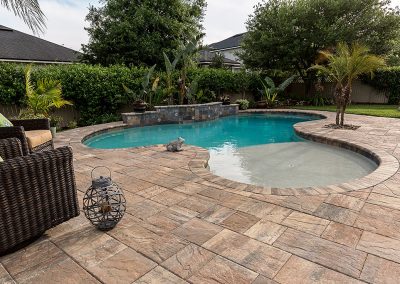 Backyard pool with stone patio, palm trees, wicker chairs, and a small raised spa in the corner.